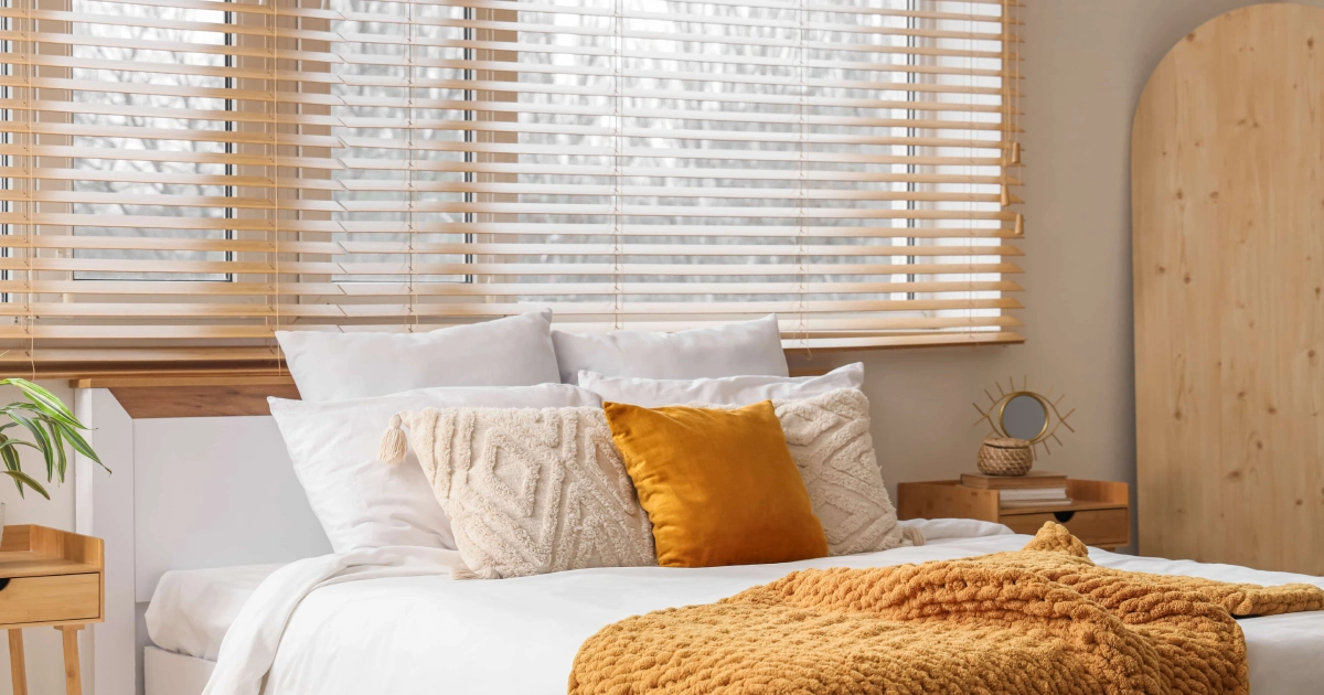 A bright bedroom with a white bed, mustard blanket, and wood blinds featuring Window Shadings in South Walton & 30A Surrounding Areas.
